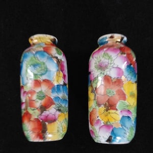 May include: A pair of small, cylindrical ceramic vases featuring a vibrant, multi-coloured floral pattern. The surface is decorated with hand-painted flowers in shades of red, blue, yellow, and pink, accented with gold trim around the narrow rims.
