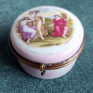 Könnte beinhalten: Kleine, runde Schmuckdose mit rosa Basis und Deckel mit einer klassischen Szene in Rosa-, Lila- und Goldtönen. Die Dose hat einen messingfarbenen Metallrand und Verschluss. Der Deckel ist mit Figuren in fließenden Gewändern bemalt.