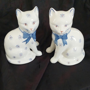 May include: Two white ceramic cat figurines featuring a hand-painted blue floral pattern across their bodies. Each cat wears a blue painted bow around its neck, with pointed ears, orange-tinted eyes, and whiskers detailed in blue paint.