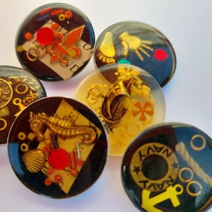 May include: Six round resin cabochons with various gold and black embellishments. The cabochons feature a variety of designs, including a fleur-de-lis, a nautical anchor, a seahorse, a coin, and a cross.