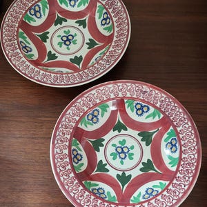 Op de afbeelding: Twee decoratieve keramische borden met een rood, wit, groen en blauw bloemmotief. De borden hebben een gedetailleerde rand en een centraal bloemmotief. De borden staan op een donker houten oppervlak.