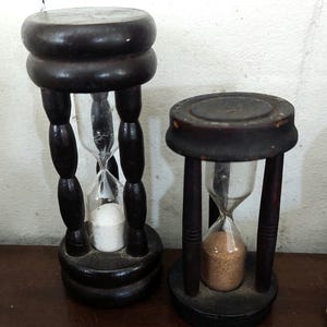 Könnte beinhalten: Zwei Sanduhren mit dunkelbraunen Holzrahmen und runden Sockeln. Die größere Sanduhr enthält weißen Sand im Glaskolben, während die kleinere Sanduhr beigen Sand enthält. Beide Rahmen verfügen über vertikale Stützen zwischen Ober- und Unterteil.