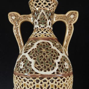 May include: A decorative, openwork ceramic vase with two handles. The vase features a cream-colored background with intricate cut-out patterns in a gold and brown color scheme. The rim and handles are accented with gold trim.