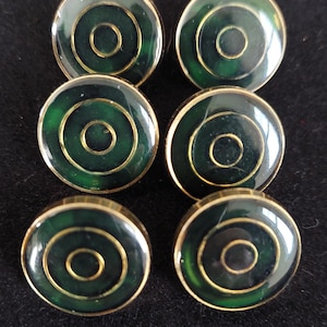 May include: Six round buttons with a dark green center and concentric gold rings. The buttons have a gold-colored rim and are arranged on a dark surface. These buttons could be used for clothing or crafts.