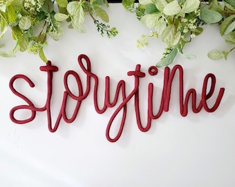 May include: A dark red knitted rope sign featuring the word storytime in a continuous cursive script. The letters are formed from a thick, textured cord that creates a soft, rounded appearance. The word is displayed in a single line of text.