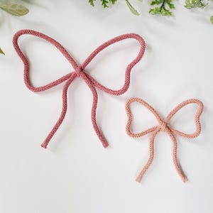 Op de afbeelding: Twee decoratieve strikken gemaakt van getextureerd touw. De grotere strik is diep oudroze van kleur, terwijl de kleinere strik een lichtere, gedempte perziktint heeft. Beide strikken zijn voorzien van een centrale knoop met twee uiteinden.