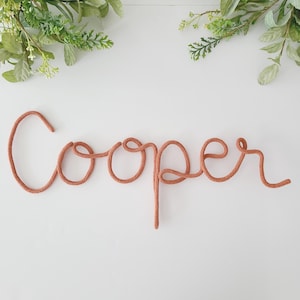 Puede incluir: Una decoración de pared con el nombre Cooper escrito en letra cursiva continua, hecha de una cuerda texturizada de color terracota. Las letras están formadas por una sola pieza de cordón, creando una apariencia suave, redondeada y flexible.