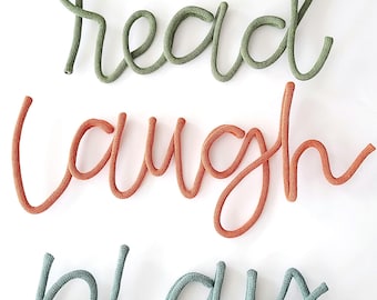 Puede incluir: Tres palabras formadas con cuerda trenzada en letra cursiva. La palabra superior es read en verde oliva, la del medio es laugh en naranja terracota y la inferior es play en azul grisáceo. Las letras están conectadas en una línea.