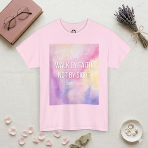May include: A light pink short-sleeve t-shirt featuring a central rectangular graphic with a watercolor wash in shades of pink, yellow, and purple. The text reads: WALK BY FAITH, NOT BY SIGHT 2 CORINTHIANS 5:7 in a clean, white, sans-serif font.
