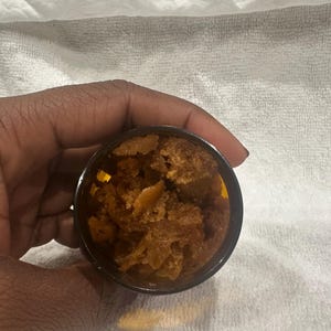 Puede incluir: Un peque&ntilde;o recipiente oscuro lleno de una sustancia granulada marr&oacute;n. El recipiente se sostiene en una mano contra un fondo blanco texturizado. El contenido parece ser un exfoliante corporal o un producto cosm&eacute;tico similar.