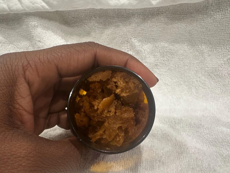 Puede incluir: Un peque&ntilde;o recipiente oscuro lleno de una sustancia granulada marr&oacute;n. El recipiente se sostiene en una mano contra un fondo blanco texturizado. El contenido parece ser un exfoliante corporal o un producto cosm&eacute;tico similar.