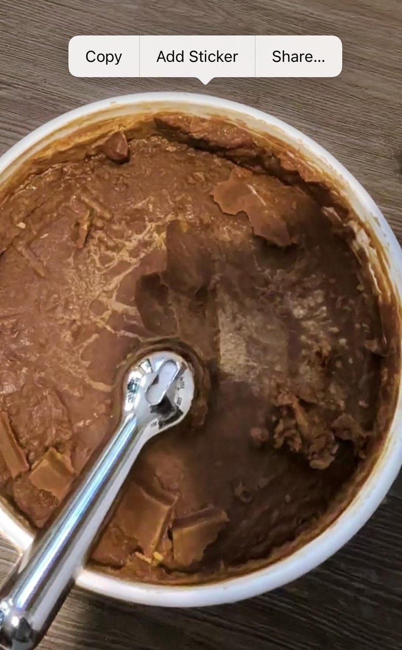 Puede incluir: Un cuenco blanco lleno de un alimento marr&oacute;n y texturizado. Una llave inglesa plateada y brillante est&aacute; parcialmente sumergida en el alimento. La comida parece tener una consistencia espesa y grumosa.