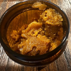 Puede incluir: Primer plano de un peque&ntilde;o frasco de vidrio &aacute;mbar oscuro lleno de una sustancia granular de color marr&oacute;n dorado. La sustancia tiene una apariencia texturizada, casi cristalina. El frasco est&aacute; sobre una superficie de madera.