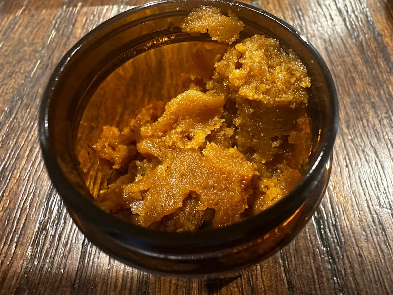 Puede incluir: Primer plano de un peque&ntilde;o frasco de vidrio &aacute;mbar oscuro lleno de una sustancia granular de color marr&oacute;n dorado. La sustancia tiene una apariencia texturizada, casi cristalina. El frasco est&aacute; sobre una superficie de madera.