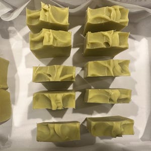 May include: Ten bars of handmade soap, each with a unique swirl pattern, are arranged in two rows on a white surface. The soap is a light green color.