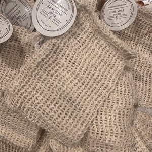 Puede incluir: Primer plano de varias bolsas de jabón de sisal. Las bolsas son de un color blanquecino natural y tienen una textura tejida en forma de red. Cada bolsa tiene una etiqueta redonda que dice "FIORI ESSENTIALS & CO" y "SISAL SOAP POUCH".