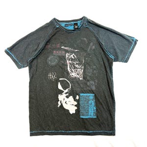 May include: Dark grey t-shirt with a crew neck and short sleeves. The shirt features a skull graphic and various text elements in white and blue. Blue stitching accents the seams and edges.