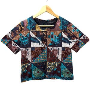 Peut inclure: Un haut court à manches courtes avec un motif géométrique dans les tons de brun, bleu et or. Le tissu présente un design patchwork avec divers motifs d'inspiration africaine.