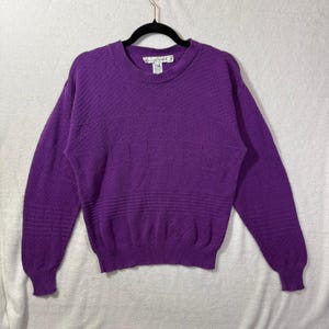 May include: A purple sweater with a crew neck and long sleeves. The sweater features a textured pattern across the chest and sleeves. The garment is hanging on a black hanger against a white background.