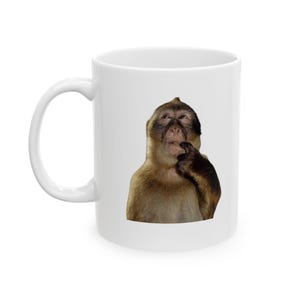 Peut inclure: Mug en céramique blanche avec un singe pensif. Le singe, avec sa fourrure brune, touche son menton. La tasse a une anse incurvée et une finition brillante. Idéale pour le café ou le thé.