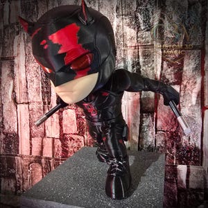 Op de afbeelding: Een zwart en rood beeldje van een superheld in een dynamische pose, met twee zilveren batons. De figuur heeft een kenmerkend masker met hoorns en rode accenten. De figuur staat op een grijs platform voor een bakstenen muur.