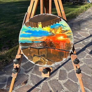 May include: A round painting on a wooden easel. The painting depicts a sunset over a cityscape with a balcony in the foreground. The painting is signed "Valentina 2021".