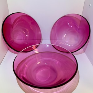 May include: Three decorative glass bowls in varying sizes. The bowls are a vibrant magenta color with a swirling pattern. Each bowl has a black rim. The bowls are displayed on a white shelf.