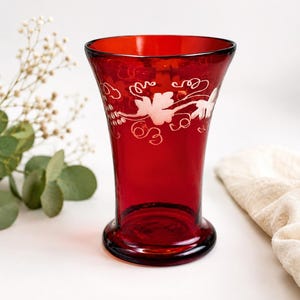 May include: A deep red glass vase featuring a flared rim and a wide base. The upper portion of the vase is decorated with an etched white pattern depicting grape leaves and vine tendrils encircling the exterior of the translucent red glass.