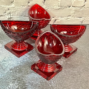 May include: Four ruby red glass dessert bowls with diamond cut patterns. Each bowl has a square base, a pedestal stem, and a rounded bowl. The bowls are arranged on a gray surface, with a white brick wall in the background.