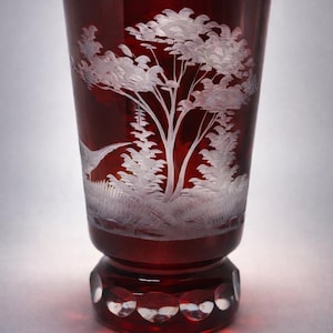 May include: A ruby red glass with a detailed white etched landscape design. The design features a tree, other foliage, and a small hill. The base of the glass has a decorative pattern.