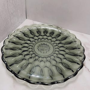 May include: A gray glass serving plate with a scalloped edge. The plate features a central floral design and repeating oval patterns around the rim. The plate is approximately 10 inches in diameter.