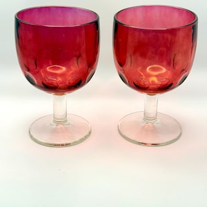 May include: Two ruby red wine glasses with clear stems and bases. The glasses have a rounded bowl shape with a textured pattern near the base of the bowl. The glasses are made of glass and are set against a white background.