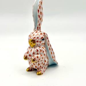 May include: A ceramic bunny figurine with a white base and a pattern of orange and brown details. The bunny's ears are long and stand upright, with a light blue interior. The bunny has gold accents on its paws and face.