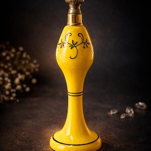 May include: A yellow ceramic perfume bottle with a decorative black floral design. The bottle has a brass-colored top and base, and is approximately 7 inches tall. The design is reminiscent of vintage apothecary bottles.