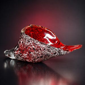 May include: A decorative glass sculpture shaped like a conch shell, featuring a vibrant red interior and an exterior surface covered in a dense, speckled pattern of black, white, and red glass fragments, with a smooth, clear glass tip at the base.