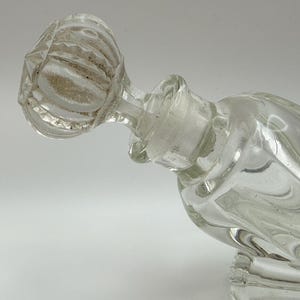 May include: Clear glass perfume bottle with a faceted stopper. The bottle has a square base and a curved body. The stopper is round with a textured, ribbed design. The bottle is empty and appears to be vintage.
