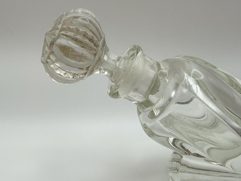 May include: Clear glass perfume bottle with a faceted stopper. The bottle has a square base and a curved body. The stopper is round with a textured, ribbed design. The bottle is empty and appears to be vintage.
