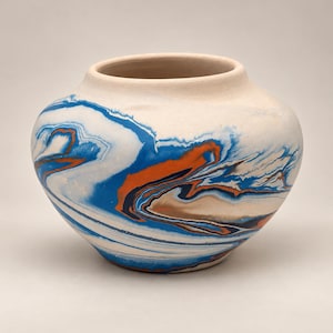 May include: A rounded ceramic vase with a wide opening and a matte finish. The exterior features a marbled pattern with swirling streaks of bright blue, burnt orange, and dark navy blue against a creamy off-white base color.