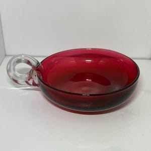 May include: A ruby red glass bowl with a clear handle. The bowl has a rounded shape and a smooth, glossy finish. The handle is circular and attached to the side of the bowl. The bowl is likely used for serving or display.