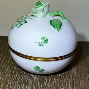 May include: A white, round trinket box with a gold band around the middle. The lid is topped with a green rose and stem. Green floral designs are painted on the white surface. A decorative piece.