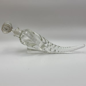 May include: Clear glass decanter with a unique, elongated design. The decanter features a textured stopper and a tail-like shape with a ridged pattern. It rests on a clear, rectangular base, showcasing its elegant form. A decorative piece for serving beverages.