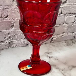 May include: A ruby red Fostoria Argus goblet. The glass features a textured design on the bowl and a round base. The goblet has a long stem and a sticker that reads "Fostoria Argus".