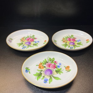 May include: Three white porcelain coasters with gold trim and floral designs. Each coaster features a different arrangement of colorful flowers with green leaves.