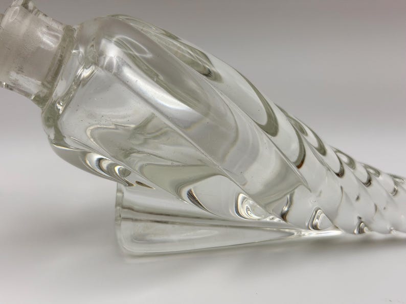 May include: Clear glass perfume bottle with a spiral design. The bottle has a long, twisted body and a small, rounded base. The stopper is visible at the top. The bottle is made of clear glass and is likely vintage.