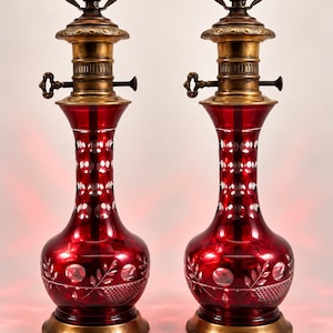 May include: A pair of table lamps featuring ruby red glass bodies with cut-glass patterns, including circular motifs and leaf designs. Each lamp has a brass base, a brass neck with a key-shaped adjustment knob, and a standard light bulb socket.