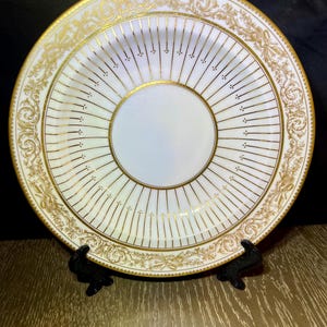 May include: White porcelain plate with gold detailing. The plate features a central white circle, surrounded by gold lines radiating outwards. The rim has ornate gold floral and scrollwork. The plate is displayed on a black stand.