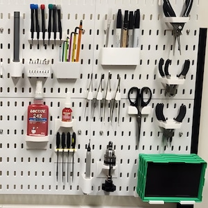 Könnte beinhalten: Eine weiße Lochwand mit verschiedenen Werkzeugen und Zubehör, darunter Stifte, Zangen, ein Hammer und ein Maßband. Die Werkzeuge sind mit weißen Haltern an der Tafel angeordnet. Eine Uhr ist ebenfalls sichtbar.