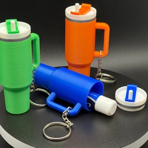May include: Three colorful, 3D-printed tumblers with handles and keychains. One is green, one is orange, and one is blue, with white and colored lids. The blue tumbler holds a tube of moisturizer. The tumblers are designed to hold a standard size beverage.
