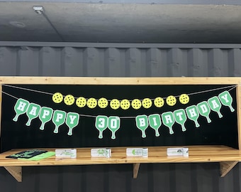 May include: A green and white banner with the words "Happy 30th Birthday" strung with yellow and black pickleball balls.