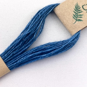 May include: A skein of deep blue embroidery thread looped through two light brown paper bands. One band features a small green fern illustration. The thread has a visible fibrous texture and is arranged in a long, continuous, folded loop.